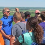 Man teaching a group of children while they view the farm country in the distance