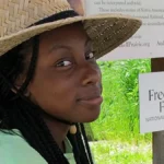 Young Black woman in a straw hat smiling gently at the camera inside the kiosk sun-shade learning area