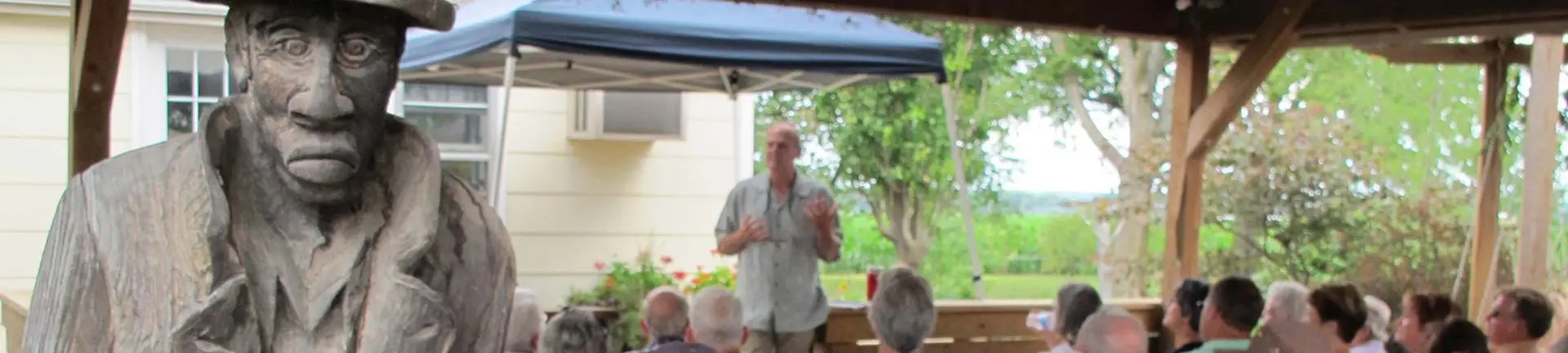 Man speaking to an audience under a shaded kiosk with wooden carved figure in the foreground